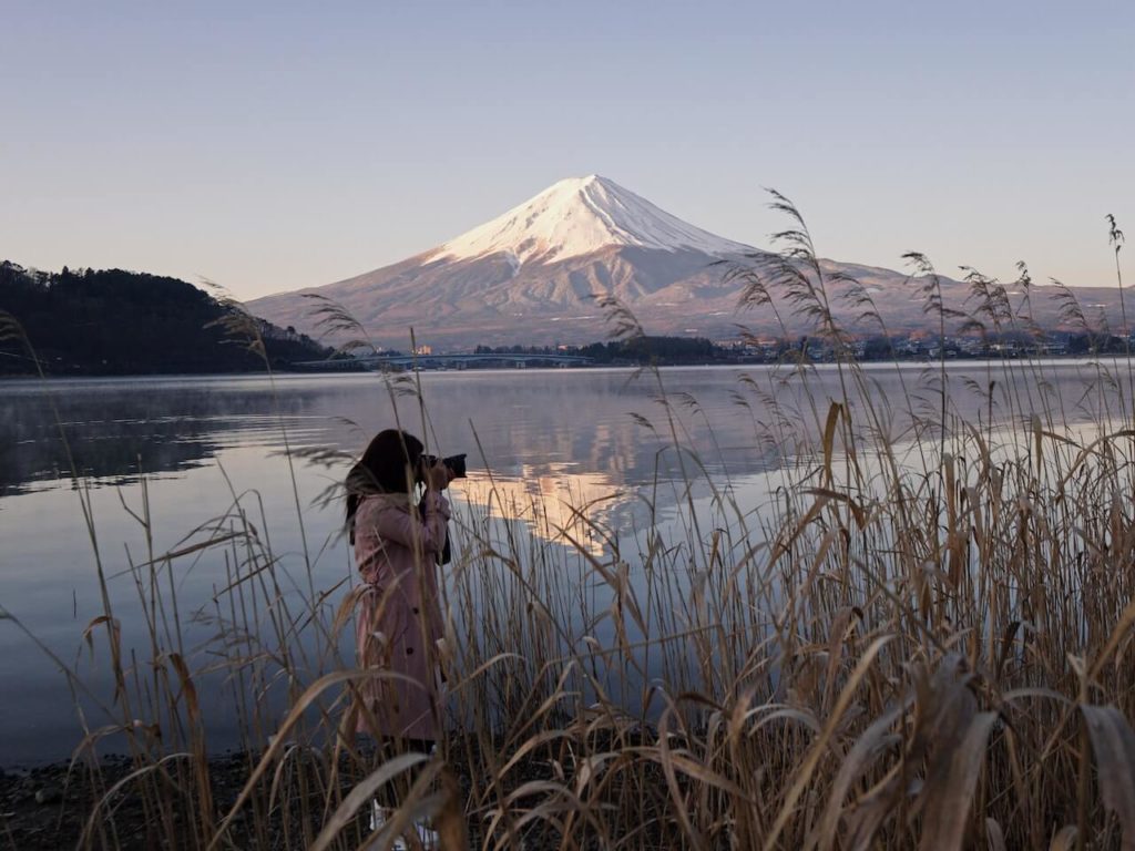 A picture of Mount Fuji, in Japan, taken from the otherside of the lake. There is a lady using a large camera infront of the Mountain.