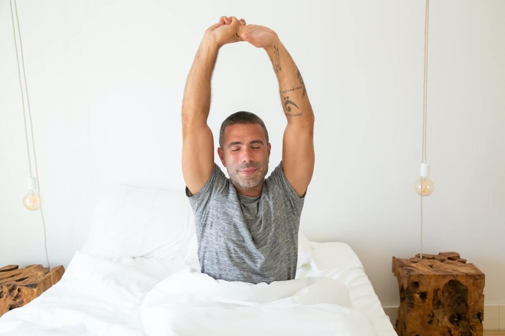 Cancel out morning depression: A man in a grey t shirt with tattoos on his left arm, sat in his bed stretching with his hands reached up over his head.